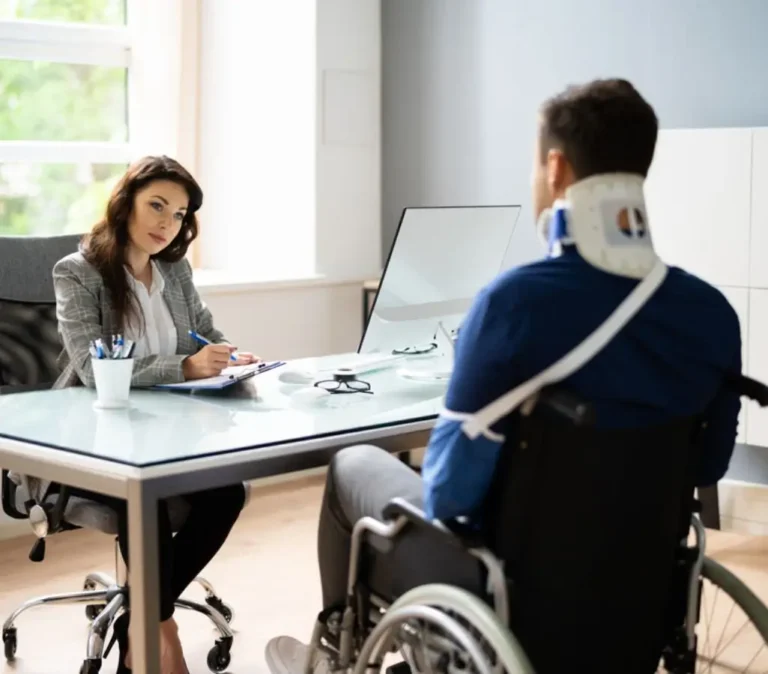 Obrazek wyróżniający do artykułu - renta z tytułu niezdolności do pracy. Na zdjęciu widzimy kobietę siedzącą za biurkiem, naprzeciwko mężczyzny na wózku inwalidzkim. Mężczyzna ma na szyi stabilizator i nosi granatową ortezę na ręce. Kobieta, może być lekarzem orzecznikiem. Notuje informacje. Scena sugeruje konsultację lub rozmowę na temat niezdolności do pracy lub świadczeń. Wzrok kobiety jest skierowany na mężczyznę, co może sugerować troskę i zaangażowanie w jego sytuację.