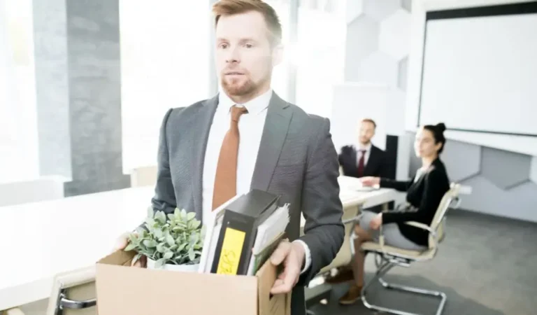 Obrazek wyróżniający w artykule - urlop na poszukiwanie pracy. Przedstawia mężczyznę w garniturze, który niesie pudło pełne rzeczy biurowych, takich jak dokumenty, roślina doniczkowa i osobiste przedmioty. Wygląda na to, że opuszcza miejsce pracy, co może wzbudzać skojarzenia z utratą pracy lub zakończeniem etapu zawodowego. Atmosfera zdjęcia wydaje się być napięta i melancholijna.