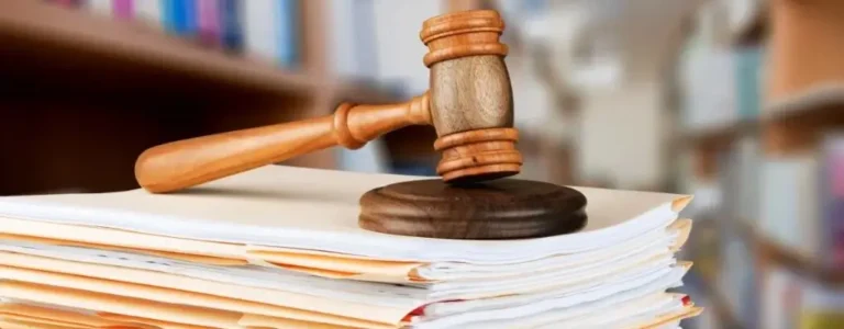 Obrazek wyróżniający w artykule - zaświadczenie o niekaralności. Na zdjęciu znajduje się drewniany młotek sędziowski umieszczony na stosie dokumentów, co jest typowym symbolem sądownictwa i praworządności. Przedstawia on decydujący moment w procesach sądowych, kiedy sędzia wydaje wyrok, kończąc tym samym rozprawę. Młotek spoczywający na dokumentach może wywoływać skojarzenia z wydaniem wyroku.