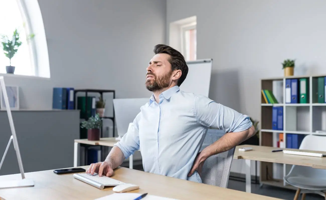 Jak otrzymać odszkodowanie za chorobę zawodową? 1 Zdjęcie główne dodane do artykułu "Sprawdź, jak uzyskać odszkodowanie za chorobę zawodową!" Na obrazku mężczyzna w niebieskiej koszuli siedzi na biurowym krześle i trzyma się za plecy z wyrazem bólu na twarzy. Wydaje się, że odczuwa dyskomfort lub ból pleców. W tle znajduje się biurowe wnętrze z biurkiem, na którym leży telefon komórkowy i klawiatura, oraz regał z segregatorami. Pomieszczenie jest jasne, z dużym oknem w dachu, przez które wpada naturalne światło.