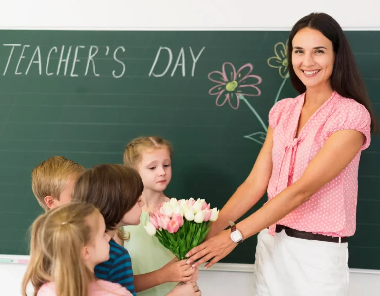 Na zdjęciu widzimy uśmiechniętą nauczycielkę o ciemnych, długich włosach, stojącą przed tablicą, na której napisano "TEACHER'S DAY" (Dzień Nauczyciela). Nauczycielka ma na sobie różową, w białe kropki bluzkę i białe spodnie. Przyjmuje bukiet tulipanów od swoich młodych uczniów. Grupa dzieci stoi odwrócona bokiem do kamery, skupiona na wręczaniu kwiatów. Cała scena rozgrywa się w jasnej, przestronnej klasie, co dodaje ciepła i przytulności obrazowi. Atmosfera jest radosna i serdeczna, a wydarzenie wydaje się być miłym gestem uznania dla pracy nauczyciela.