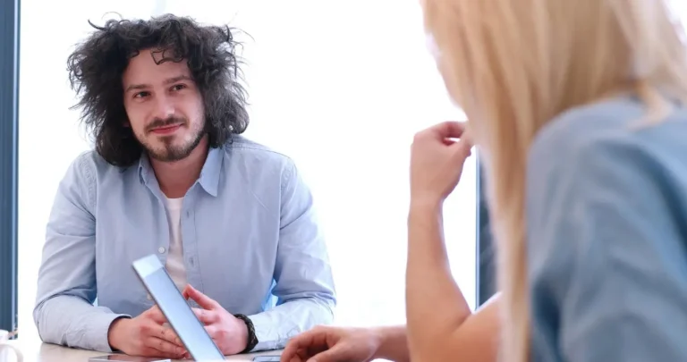 Na zdjęciu widzimy młodego mężczyznę z kręconymi, czarnymi włosami, który ma na sobie jasnoniebieską koszulę. Siedzi naprzeciwko nieco niewyraźnej postaci kobiety z blond włosami. Mężczyzna uśmiecha się delikatnie i wydaje się prowadzić rozmowę, patrząc bezpośrednio na kobietę. Jego postawa jest otwarta i zrelaksowana, a ręce ma splecione na stole. Scena ta sugeruje, że może to być profesjonalne spotkanie, być może rozmowa kwalifikacyjna lub konsultacja, w jasnym i nowoczesnym biurze. Tło jest jasne, co dodaje atmosferze spokoju i profesjonalizmu.