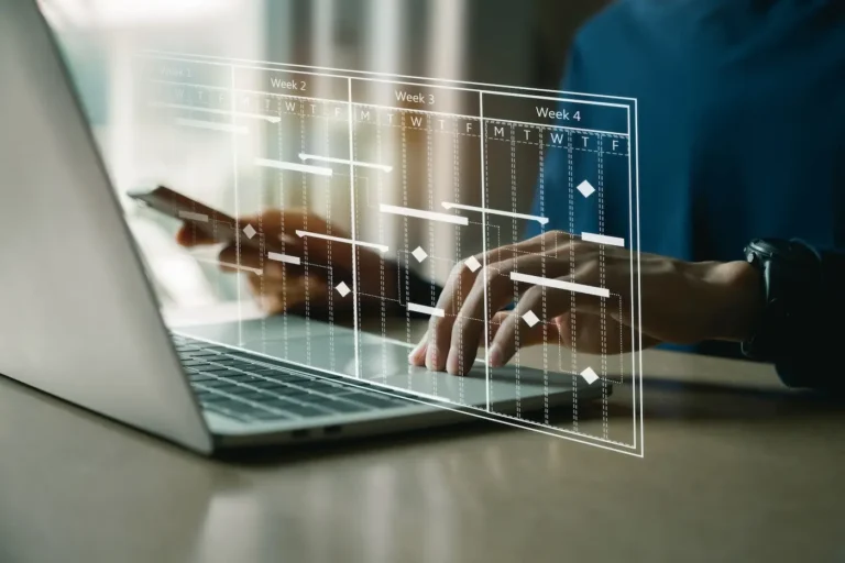 Zdjęcie przedstawia osobę pracującą na laptopie, nad którą wyświetlają się holograficzne obrazy interfejsu z harmonogramem lub kalendarzem z podziałem na tygodnie. Scena sugeruje nowoczesne zarządzanie czasem lub planowanie w biznesie przy użyciu zaawansowanych technologicznie narzędzi. Osoba ma na nadgarstku czarny zegarek, co może wskazywać na profesjonalizm i dbałość o terminowość.