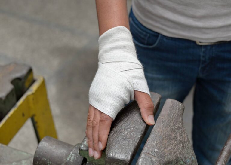 Na zdjęciu widoczna jest osoba z bandażowaną ręką, która opiera dłoń o metalowy przedmiot, wyglądający na część jakiejś maszyny lub urządzenia. Bandaż jest śnieżnobiały i owinął większość dłoni oraz nadgarstka, sugerując niedawny uraz lub zabieg medyczny. Osoba ma na sobie szary T-shirt oraz niebieskie jeansy, co może wskazywać na nieformalny strój roboczy. Tło jest nieostre, ale wygląda na przemysłowe lub warsztatowe. Scena wydaje się podkreślać znaczenie bezpieczeństwa i ochrony zdrowia w miejscu pracy.