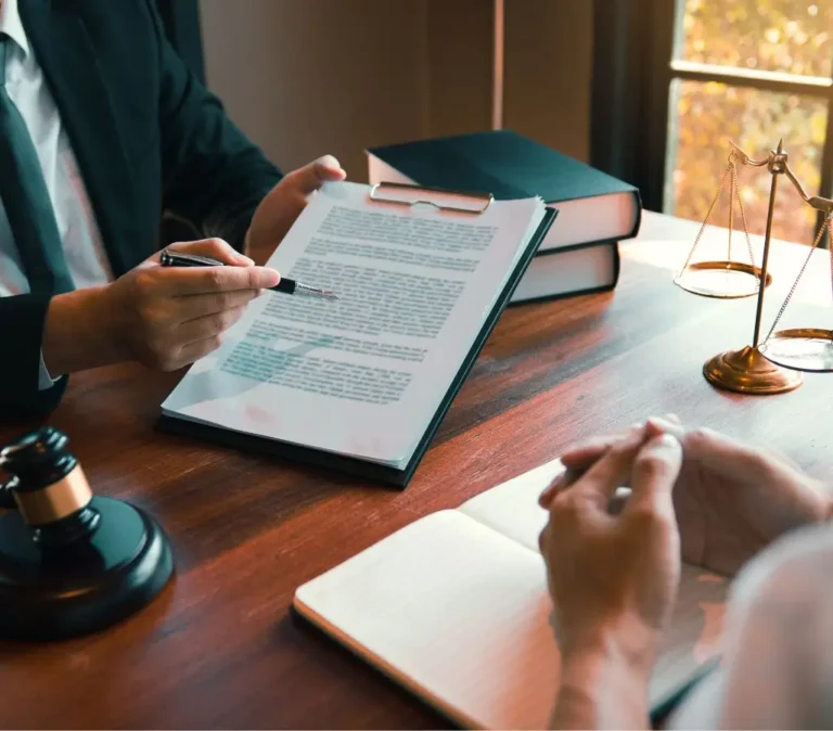 Obrazek wyróżniający w artykule - Darmowa pomoc prawna Warszawa. Na zdjęciu widać osobę będącą prawnikiem lub doradcą prawnym, która pokazuje palcem fragment tekstu na dokumencie. Osoba ta jest ubrana w ciemną marynarkę i trzyma długopis. Naprzeciwko niej, po drugiej stronie biurka, siedzi klient lub klientka z założonymi rękoma, których twarz nie jest widoczna. Na biurku po prawej stronie znajduje się mosiężna waga – symbol sprawiedliwości. W tle na biurku leżą zamknięte książki prawne oraz notatnik. Całość wygląda na scenę z kancelarii prawniczej, a na zewnątrz przez okno widać drzewa, co sugeruje, że biuro mieści się na parterze lub pierwszym piętrze.