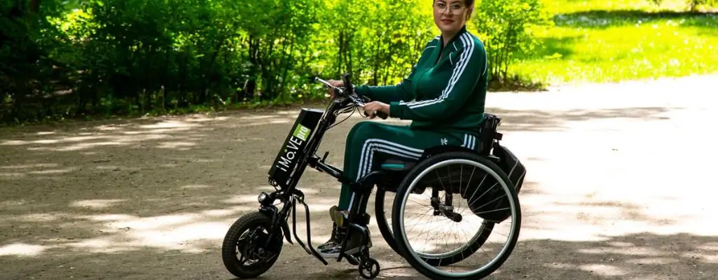 Zdjęcie w artykule - dofinansowanie na przystawkę elektryczną do wózka. Kobieta w wózku inwalidzkim z zamocowaną zieloną elektryczną przystawką, poruszająca się po ścieżce w parku. Kobieta ma na sobie sportowy strój i wygląda na zadowoloną, co sugeruje, że dzięki przystawce może cieszyć się większą swobodą poruszania i aktywnością na świeżym powietrzu. Scena rozgrywa się na tle leśnej ścieżki i zielonej roślinności w słoneczny dzień. Widać, że kobieta trzyma kierownicę napędu, przygotowując się do jazdy.