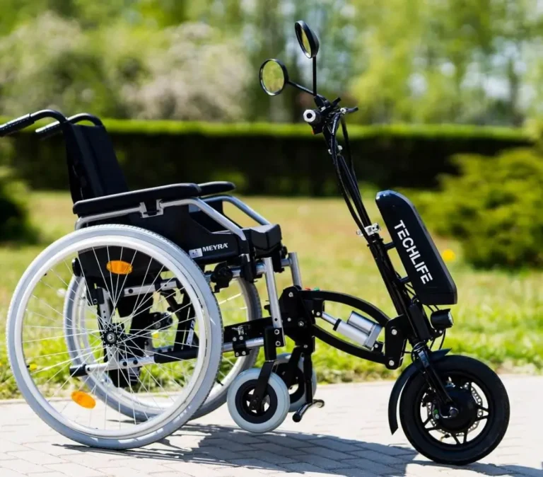 Obrazek wyróżniający do artykułu - dofinansowanie na przystawkę elektryczną do wózka. Zdjęcie pokazuje wózek inwalidzki ręczny, do którego przyczepiony jest czarny, elektryczny napęd z napisem "TECHLIFE". Napęd wyposażony jest w małe, czarne koło oraz lusterko wsteczne. Całość znajduje się na tle zielonego ogrodu z drzewami i krzewami. Jest to urządzenie, które ma na celu ułatwienie i zwiększenie mobilności osób korzystających z wózka inwalidzkiego.