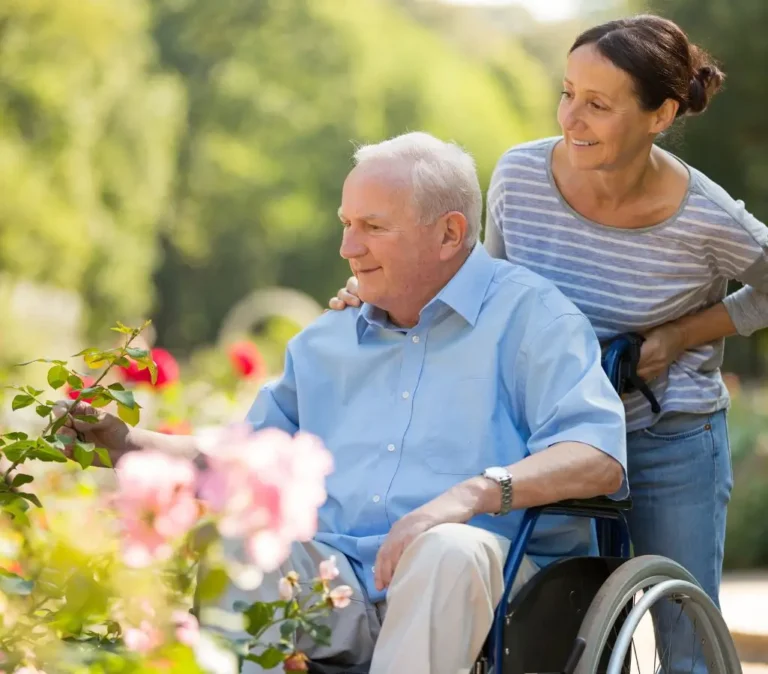 Obrazek wyróżniający do artykułu - koniec świadczenia pielęgnacyjnego co dalej. Na zdjęciu widzimy starszego mężczyznę na wózku inwalidzkim i opiekunkę. Mężczyzna wydaje się być zadowolony, patrzy na kwiaty, do których wyciąga rękę. Kobieta wygląda na uśmiechniętą i skupioną na mężczyźnie. Otoczenie jest zielone i słoneczne, prawdopodobnie są w parku lub ogrodzie.