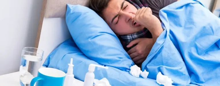 Obrazek wyróżniający do artykułu - na ile dni opłaca się brać zwolnienie lekarskie. Zdjęcie ukazuje młodą osobę leżącą w łóżku, okrytą niebieską kołdrą. Osoba ta wygląda na chorą, ma na sobie szalik i trzyma się za gardło, co może sugerować ból czy dyskomfort. Na nocnym stoliku obok łóżka widać wodę, leki, chusteczki i inne przedmioty, które zwykle towarzyszą przeziębieniu czy grypie. Obraz ten może odzwierciedlać uczucie zmęczenia oraz potrzebę odpoczynku i leczenia.