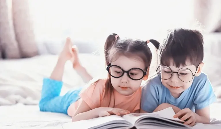 obrazek wyróżniający do artykułu okulary do czytania. Zdjęcie przedstawia dwoje małych dzieci leżących na brzuchu, skupionych na czytaniu dużej otwartej książki. Dzieci mają na sobie okulary, które dodatkowo podkreślają ich zaangażowanie w czytanie. Obrazek sugeruje, że okularów do czytania potrzebują osoby w różnym wieku.