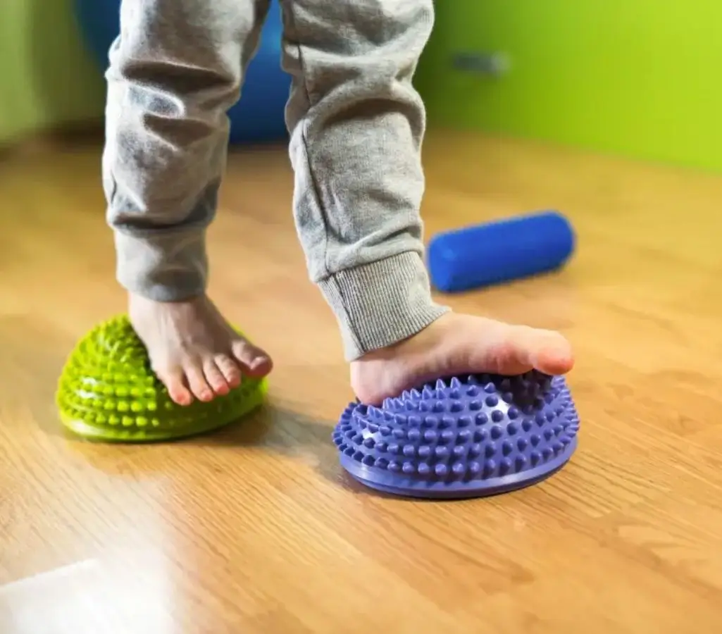 Płaskostopie u dzieci od A do Z. Wszystko, co warto wiedzieć o tej wadzie stóp 1 Obrazek wyróżniający w artykule płaskostopie u dzieci. Na zdjęciu widać stopy dziecka stojącego na kolorowych, wypukłych poduszkach sensorycznych - jedna jest zielona, a druga niebieska. Takie poduszki są często używane w celu stymulacji sensorycznej, poprawy koordynacji oraz wzmacniania mięśni stóp, co może być elementem terapii dzieci z płaskostopiem lub innymi dysfunkcjami stóp. Dziecko ma na sobie szare dresowe spodnie, a tło zdjęcia to drewniana podłoga oraz jasnozielona ściana, co sugeruje, że scena rozgrywa się w przestrzeni domowej lub terapeutycznej.