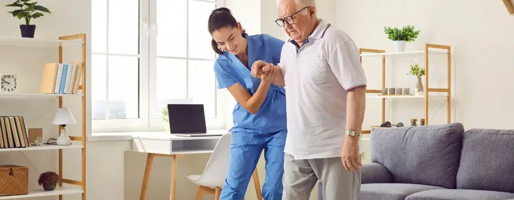 Zdjęcie w artykule - Rehabilitacja domowa NFZ. Zdjęcie przedstawia wnętrze jasnego pokoju, w którym starszy mężczyzna, ubrany w biały t-shirt z krótkim rękawem i szare spodnie, otrzymuje pomoc od młodej kobiety w niebieskim uniformie medycznym. Kobieta, prawdopodobnie rehabilitantka, uśmiecha się i wspiera mężczyznę, trzymając go pod rękę i pomagając mu w chodzeniu. W tle widać biurko z laptopem, półki z książkami i roślinami oraz kanapę. Sceneria sugeruje, że to rehabilitacja odbywa się w domowym otoczeniu pacjenta.