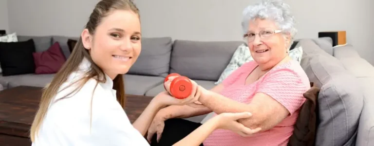 Obrazek wyróżniający do artykułu - Rehabilitacja domowa NFZ. Na zdjęciu widzimy starszą panią z siwymi włosami, ubraną w różową bluzkę, która wykonuje ćwiczenia rehabilitacyjne z pomocą młodej kobiety w białym uniformie. Starsza pani trzyma w prawej dłoni czerwoną hantlę i wygląda na to, że jest w trakcie podnoszenia jej z pomocą rehabilitantki. Siedzą one obok siebie, a scena rozgrywa się w domowym salonie, gdzie w tle widać sofę i poduszki. Panuje atmosfera wsparcia i pozytywnej interakcji pomiędzy pacjentką a terapeutką.