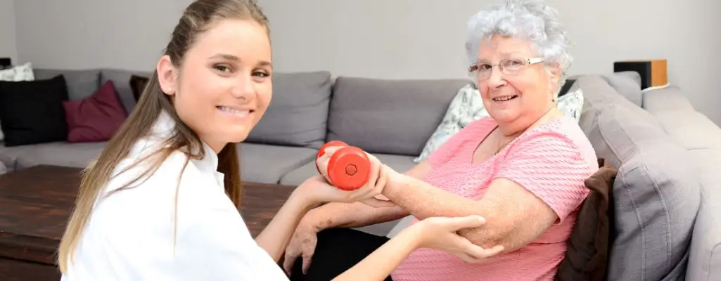 Obrazek wyróżniający do artykułu - Rehabilitacja domowa NFZ. Na zdjęciu widzimy starszą panią z siwymi włosami, ubraną w różową bluzkę, która wykonuje ćwiczenia rehabilitacyjne z pomocą młodej kobiety w białym uniformie. Starsza pani trzyma w prawej dłoni czerwoną hantlę i wygląda na to, że jest w trakcie podnoszenia jej z pomocą rehabilitantki. Siedzą one obok siebie, a scena rozgrywa się w domowym salonie, gdzie w tle widać sofę i poduszki. Panuje atmosfera wsparcia i pozytywnej interakcji pomiędzy pacjentką a terapeutką.