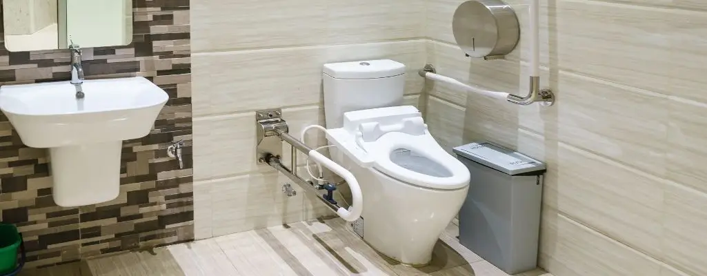 Zdjęcie w artykule - wc dla niepełnosprawnych. Na zdjęciu widoczna jest toaleta przystosowana do potrzeb osób niepełnosprawnych. W centralnym punkcie znajduje się biała muszla klozetowa z otwartą, specjalnie wyprofilowaną deską, ułatwiającą korzystanie. Po lewej stronie toalety umieszczono białą umywalkę wiszącą na ścianie, a pod nią zielony kosz na śmieci. Toaleta wyposażona jest w metalowe poręcze wspierające po obu stronach, z dodatkowym uchwytem poręczowym z przodu, co ma zapewnić bezpieczeństwo i niezależność użytkownikom. Po prawej stronie znajduje się metalowy uchwyt na papier toaletowy oraz szara metalowa kosz na odpady, stojąca na podłodze. Ściany są wyłożone jasnymi płytkami ceramicznymi z poziomym pasem dekoracyjnym z ciemniejszych płytek w środkowej części. Podłoga pokryta jest płytkami w jasnym kolorze, co współgra z ogólnym wystrojem łazienki. Ogólnie, aranżacja toalety skupia się na funkcjonalności i dostępności, z wyraźnym uwzględnieniem potrzeb osób z ograniczeniami ruchowymi.