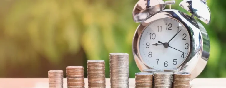 Obrazek wyróżniający do artykułu – zasiłki. Na obrazku widzimy rosnące stosy monet ułożone obok siebie na wzór wykresu słupkowego. W tle, rozmyte, znajduje się analogowy budzik. Kompozycja ta może sugerować wzrost oszczędności w czasie, symbolizując planowanie i czas na skorzystanie z odpowiedniego rodzaju zasiłku.