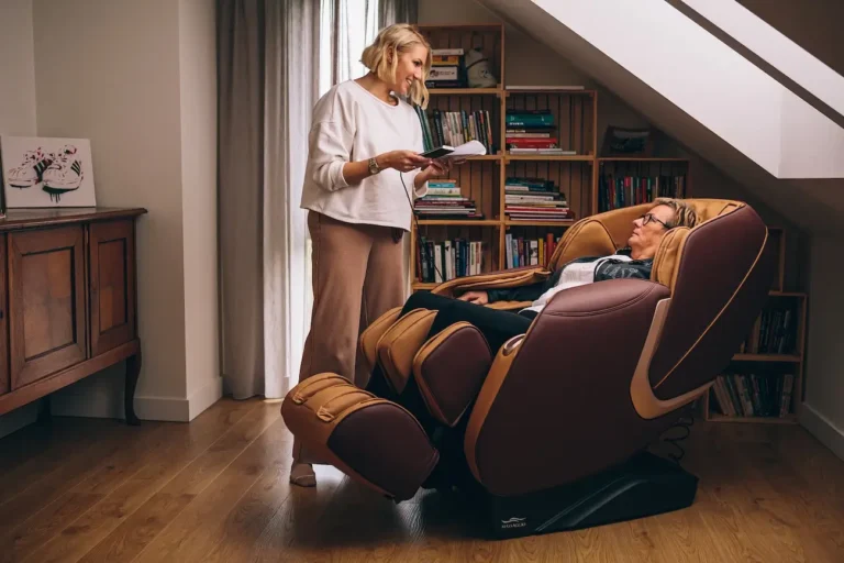 Zdjęcie przedstawia scenę w przytulnym pokoju, gdzie dwie kobiety spędzają czas. Jedna z nich, starsza kobieta, siedzi w dużym, wygodnym fotelu masującym w kolorze brązowym z elementami beżowymi. Wygląda na zrelaksowaną, ma zamknięte oczy i na twarzy wyraz odprężenia. Obok niej stoi młodsza kobieta z krótkimi blond włosami, ubrana w białą bluzę i beżowe spodnie. Trzyma w rękach notatnik i coś czyta z uśmiechem na twarzy, zwracając się do siedzącej kobiety. W tle widać regały wypełnione książkami oraz drewnianą komodę, na której stoi obrazek przedstawiający parę butów sportowych. Pokój jest dobrze oświetlony dzięki dużemu oknu z zasłoną, przez które wpada naturalne światło.