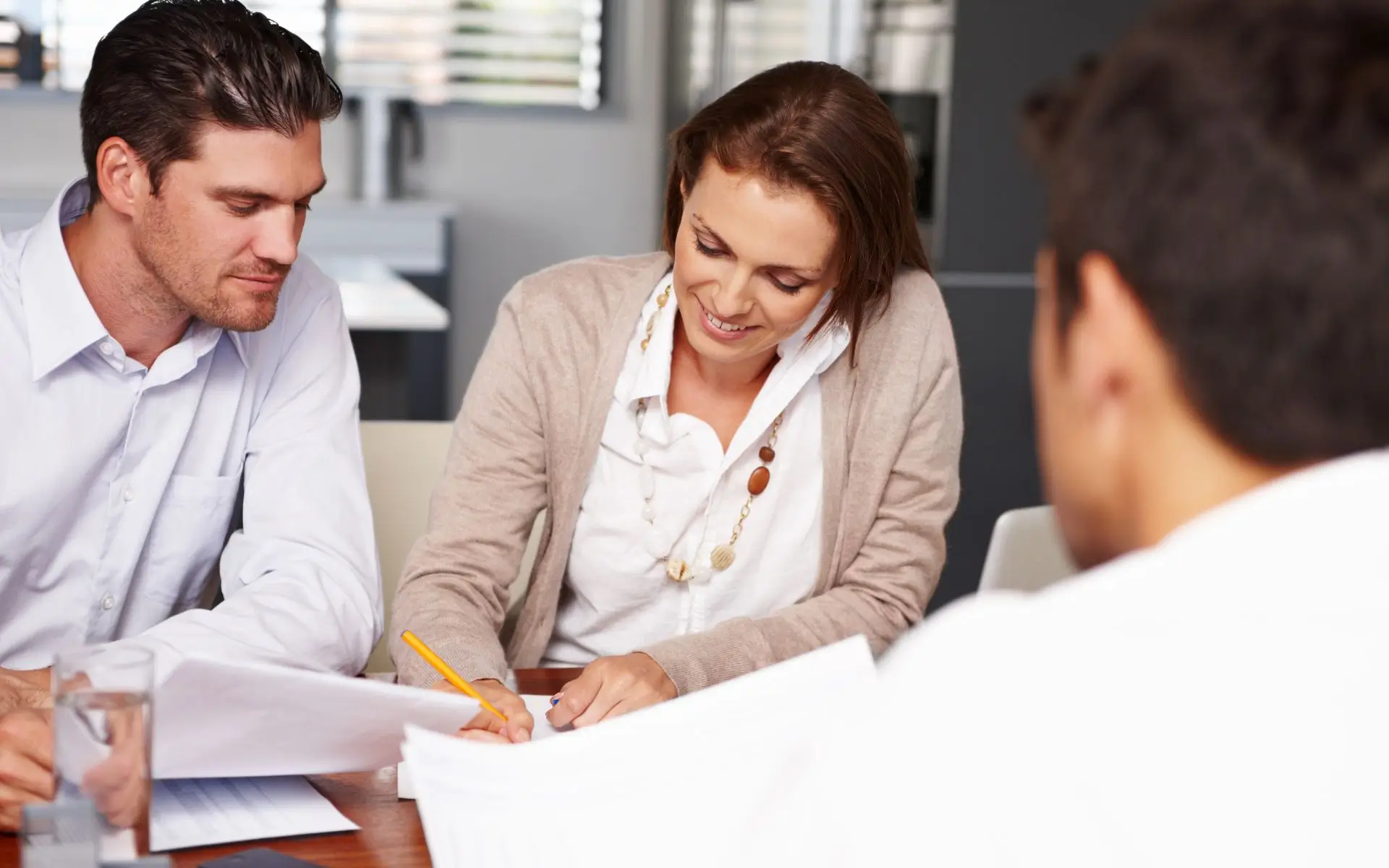 Zdjęcie przedstawia trzy osoby siedzące przy stole podczas spotkania biznesowego lub rekrutacyjnego. Dwóch mężczyzn i jedna kobieta są zaangażowani w przeglądanie i podpisywanie dokumentów. • Kobieta w środku nosi białą koszulę i beżowy sweter. Ma krótko przycięte, brązowe włosy i naszyjnik z dużymi koralikami. Uśmiecha się lekko, skupiając się na pisaniu. • Mężczyzna po lewej stronie ma krótkie, ciemne włosy i białą koszulę. Wydaje się uważnie przeglądać dokumenty. • Trzecia osoba, widoczna od tyłu, siedzi po prawej stronie. Jego twarz nie jest widoczna, ale widać ciemne włosy i białą koszulę. Na stole znajdują się dokumenty oraz szklanka wody. W tle widać niewyraźnie wnętrze biura, z oknami z żaluzjami i częściowo widocznymi meblami biurowymi.