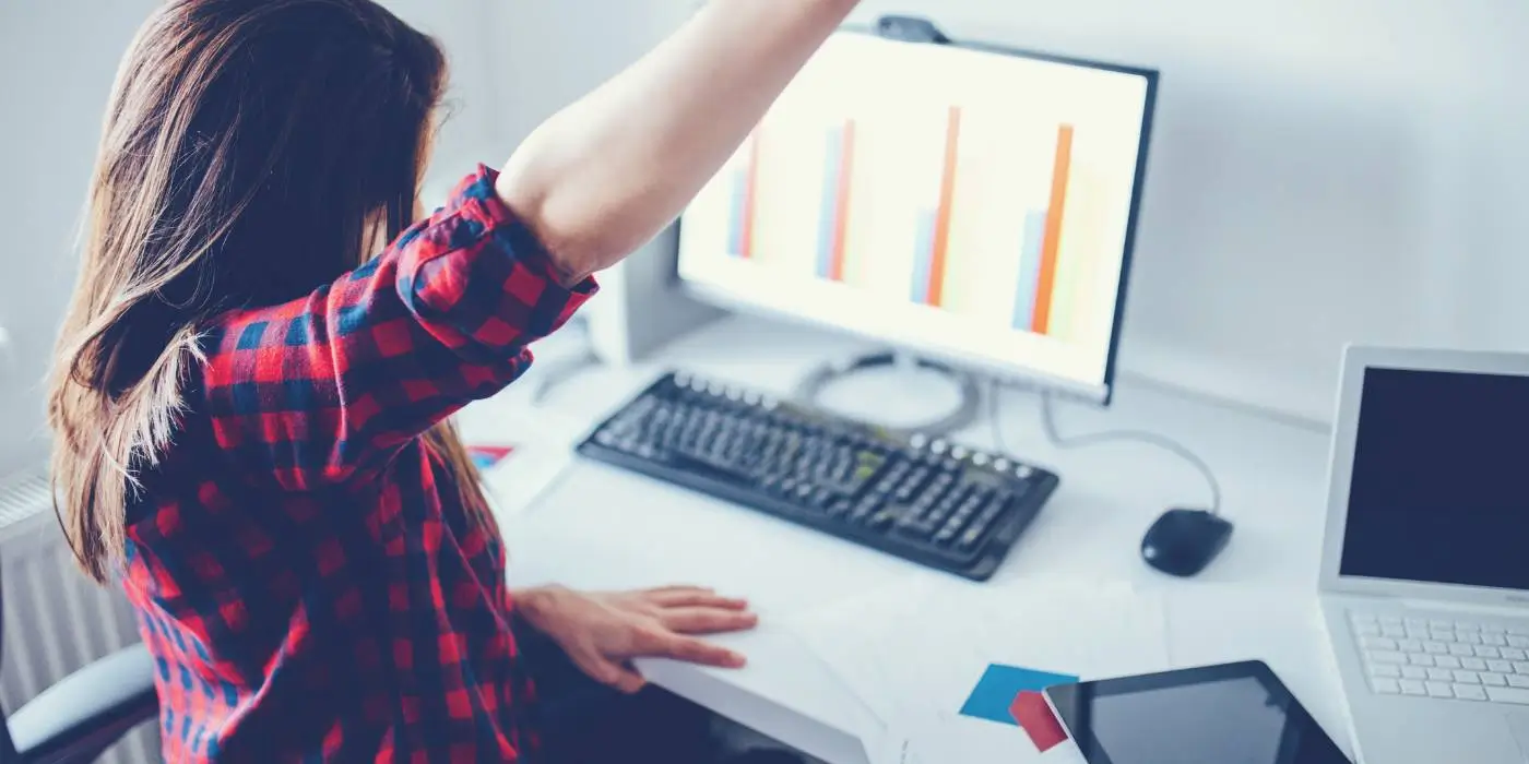 Realizacja prac B+R jako kluczowy element projektów ścieżki SMART 1.1 w ramach dotacji FENG 1 Zdjęcie przedstawia młodą kobietę w biurowym środowisku, skupioną na pracy przy komputerze. Kobieta ma długie brązowe włosy i jest ubrana w czerwono-niebieską, kratowaną koszulę. Siedzi bokiem do kamery, z uniesioną prawą ręką, co może sugerować gest rozciągania lub odpoczynek od pracy. Przed nią na biurku widoczne są różne przedmioty biurowe, takie jak klawiatura, mysz komputerowa, wykresy na papierze oraz dodatkowy monitor. Tło jest jasne i neutralne, skoncentrowane na pracy biurowej z użyciem nowoczesnej technologii. Atmosfera zdjęcia oddaje codzienne życie biurowe, z naciskiem na pracę przy danych i analizach.