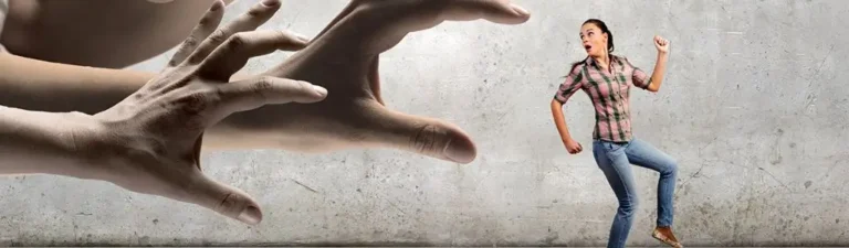 Obrazek wyróżniający w artykule - jak udowodnić mobbing w pracy. Na obrazie widzimy młodą kobietę w dżinsach i w kratowaną koszulę, która wygląda na przerażoną i uciekającą przed dwiema dużymi, nadciągającymi dłońmi. Dłonie te, które są nieproporcjonalnie większe niż postać kobiety, wydają się sięgać w jej kierunku w agresywny sposób. Cała scena rozgrywa się na tle surowej, betonowej ściany, co dodaje atmosfery zagrożenia i bezosobowości. Obraz ten może symbolizować uczucie zagrożenia, strachu przed dominacją lub kontrolą, co jest często odczuwalne przez ofiary zastraszania lub mobbingu w miejscu pracy.