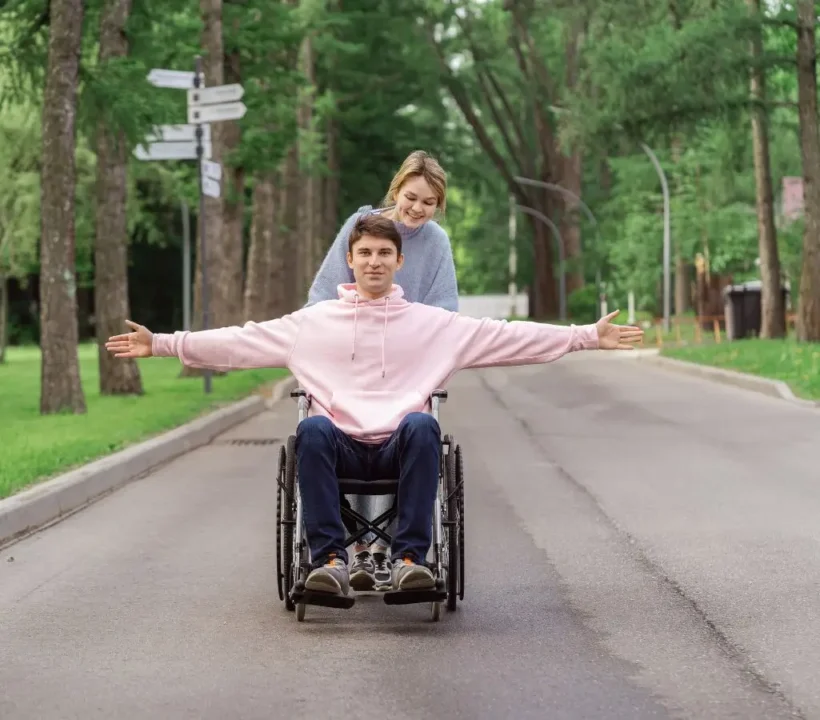 Obrazek wyróżniający do artykułu - jak zachowywać się wobec osób niepełnosprawnych. Na zdjęciu widać młodego mężczyznę na wózku inwalidzkim i kobietę za nim, która popycha wózek. Mężczyzna w różowej bluzie z kapturem ma rozłożone ręce na boki, co sugeruje radość i poczucie wolności. Kobieta, ubrana w szarą bluzę, uśmiecha się i wygląda na szczęśliwą, towarzysząc mężczyźnie. Są na ścieżce w parku, otoczeni zielenią i drzewami. Scena wyraża ciepło, współpracę i przyjemność ze wspólnie spędzanego czasu na łonie natury.