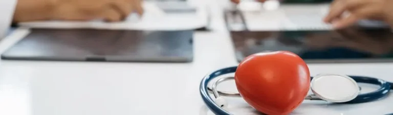 Obrazek wyróżniający do artykułu - sanatorium dla osób ze znacznym stopniem niepełnosprawności. Na zdjęciu widać fragment biurka z lekarskimi narzędziami, na którym położony jest stetoskop oraz czerwone serce symbolizujące opiekę zdrowotną. W tle rozmyte są ręce osób pracujących na tabletach lub komputerach, co sugeruje nowoczesne podejście do opieki zdrowotnej z użyciem technologii. Całość tworzy obraz zajętych, ale zaangażowanych profesjonalistów medycznych, którzy korzystają z nowoczesnych narzędzi w celu zapewnienia najlepszej możliwej opieki pacjentom.