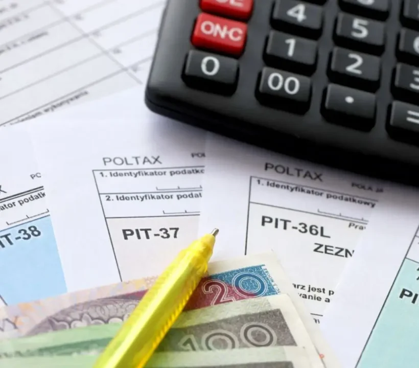 Obrazek wyróżniający do artykułu - ulga na dziecko pełnoletnie. Na zdjęciu widoczne są formularze podatkowe oznaczone jako "POLTAX" z różnymi numerami PIT, w tym PIT-37 i PIT-36L. Na dokumentach leży żółty długopis oraz polskie banknoty o nominałach 20 i 200 złotych, a tuż obok dokumentów znajduje się kalkulator. Scena ta oddaje atmosferę skupienia i porządku związaną z przygotowaniem deklaracji podatkowych, podkreślając ważność i precyzję w zarządzaniu finansami. Zdjęcie może przywoływać uczucia odpowiedzialności i powagi, jakie niosą za sobą obowiązki podatkowe, a także ulgę wynikającą z możliwości korzystania z ulg, takich jak ulga na dziecko pełnoletnie.