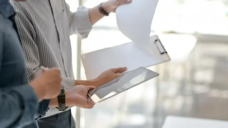 Na zdjęciu widać dwie osoby, które przeglądają dokumenty. Jedna osoba trzyma w rękach tablet, na którym prawdopodobnie wyświetlane są jakieś informacje, a druga osoba trzyma teczkę z dokumentami i przegląda kartki. Obie osoby są ubrane w koszule, co sugeruje, że znajdują się w środowisku biznesowym lub biurowym. Tło jest rozmyte, ale wydaje się, że zdjęcie zostało zrobione w jasnym, dobrze oświetlonym pomieszczeniu z dużą ilością naturalnego światła.