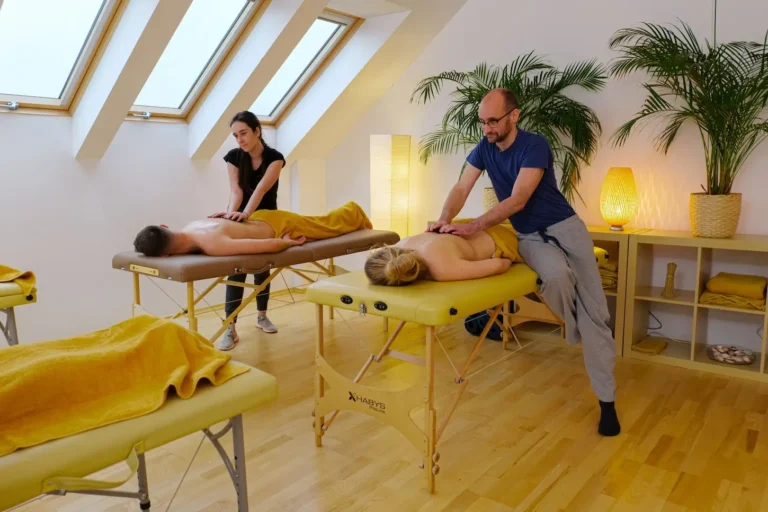 Na zdjęciu znajduje się sala do masażu z dwoma łóżkami do masażu ustawionymi równolegle obok siebie. Po lewej stronie młoda kobieta o ciemnych włosach, ubrana w czarną koszulkę i spodnie, masuje plecy mężczyzny leżącego na brzuchu. Po prawej stronie mężczyzna z krótkimi włosami i brodą, ubrany w niebieską koszulkę i szare spodnie, masuje plecy kobiety również leżącej na brzuchu. Pomieszczenie jest jasne, z dużymi oknami dachowymi, które wpuszczają dużo naturalnego światła. Na ścianie w tle znajdują się rośliny w doniczkach i lampa, tworząc przytulną i relaksującą atmosferę. Oba łóżka do masażu są pokryte żółtymi ręcznikami. Podłoga jest drewniana, co dodaje ciepła do wystroju wnętrza.