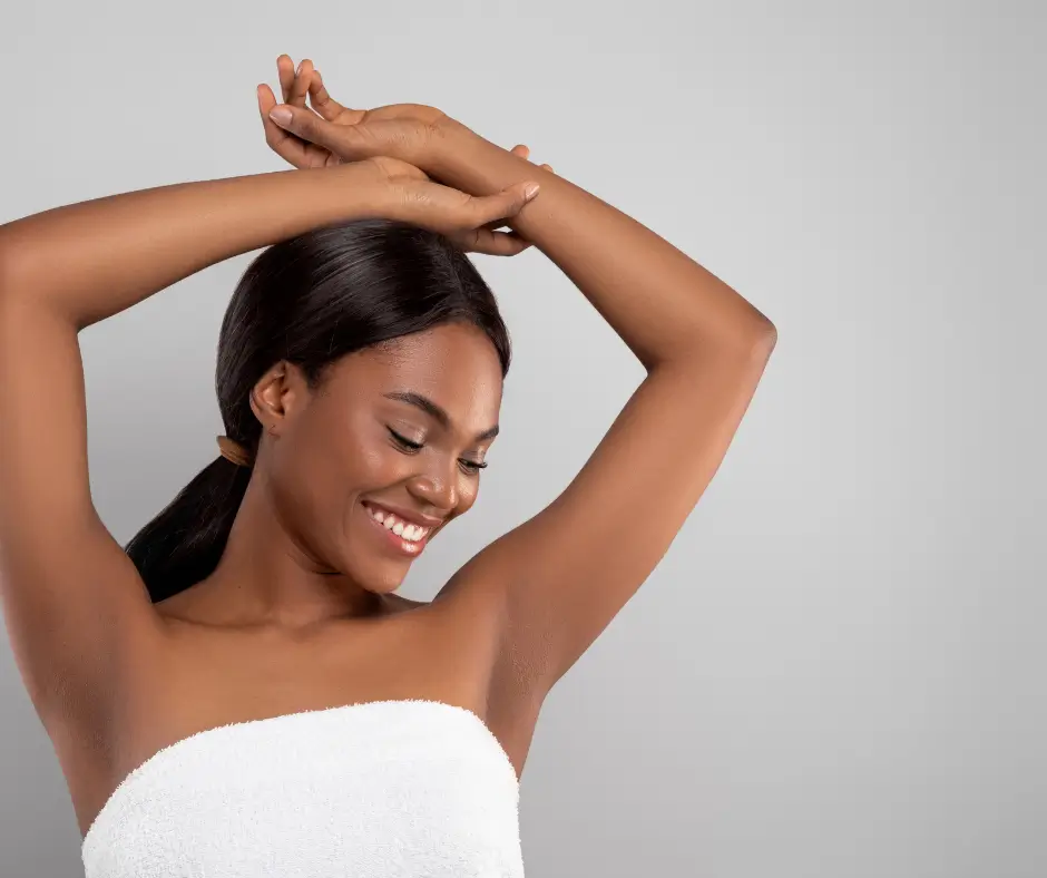 Analizujemy, jakie metody depilacji będą dla Ciebie odpowiednie 1 Na zdjęciu widzimy młodą kobietę o ciemnej karnacji i gładkiej skórze, ubraną w biały ręcznik. Kobieta ma związane włosy, a jej twarz wyraża radość, ponieważ uśmiecha się szeroko, spoglądając w dół. Jej ręce są uniesione nad głową i skrzyżowane, co eksponuje gładkie pachy, sugerujące, że skóra została świeżo wydepilowana. Tło zdjęcia jest jednolite, szare, co sprawia, że postać kobiety jest wyraźnie widoczna. Fotografię można skojarzyć z reklamą produktów do depilacji lub kosmetyków pielęgnacyjnych.