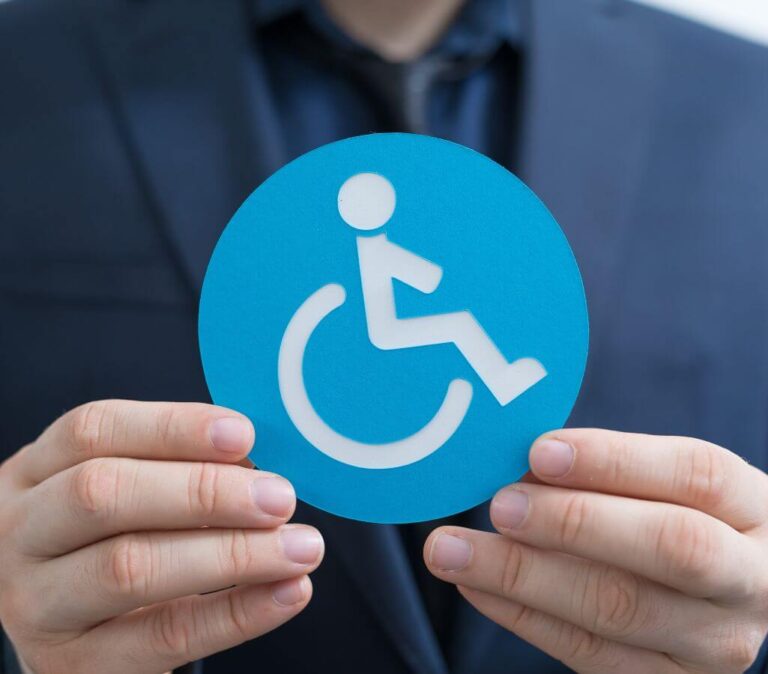 Obrazek wyróżniający do artykułu - ulgi dla niepełnosprawnych. Zdjęcie przedstawia osobę w garniturze trzymającą niebieski znak z symbolem osoby na wózku inwalidzkim, symbolizujący niepełnosprawność. Znajduje się on w centrum kadru i jest dobrze widoczny na tle ciemniejszego ubioru osoby, co sprawia, że znak wyraźnie się wyróżnia. Symbol ten jest powszechnie rozpoznawany i kojarzony z udogodnieniami oraz prawami dla osób z niepełnosprawnościami.