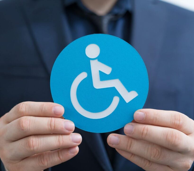 Sprawdzamy, na jakie ulgi dla niepełnosprawnych możesz liczyć 3 Obrazek wyróżniający do artykułu - ulgi dla niepełnosprawnych. Zdjęcie przedstawia osobę w garniturze trzymającą niebieski znak z symbolem osoby na wózku inwalidzkim, symbolizujący niepełnosprawność. Znajduje się on w centrum kadru i jest dobrze widoczny na tle ciemniejszego ubioru osoby, co sprawia, że znak wyraźnie się wyróżnia. Symbol ten jest powszechnie rozpoznawany i kojarzony z udogodnieniami oraz prawami dla osób z niepełnosprawnościami.