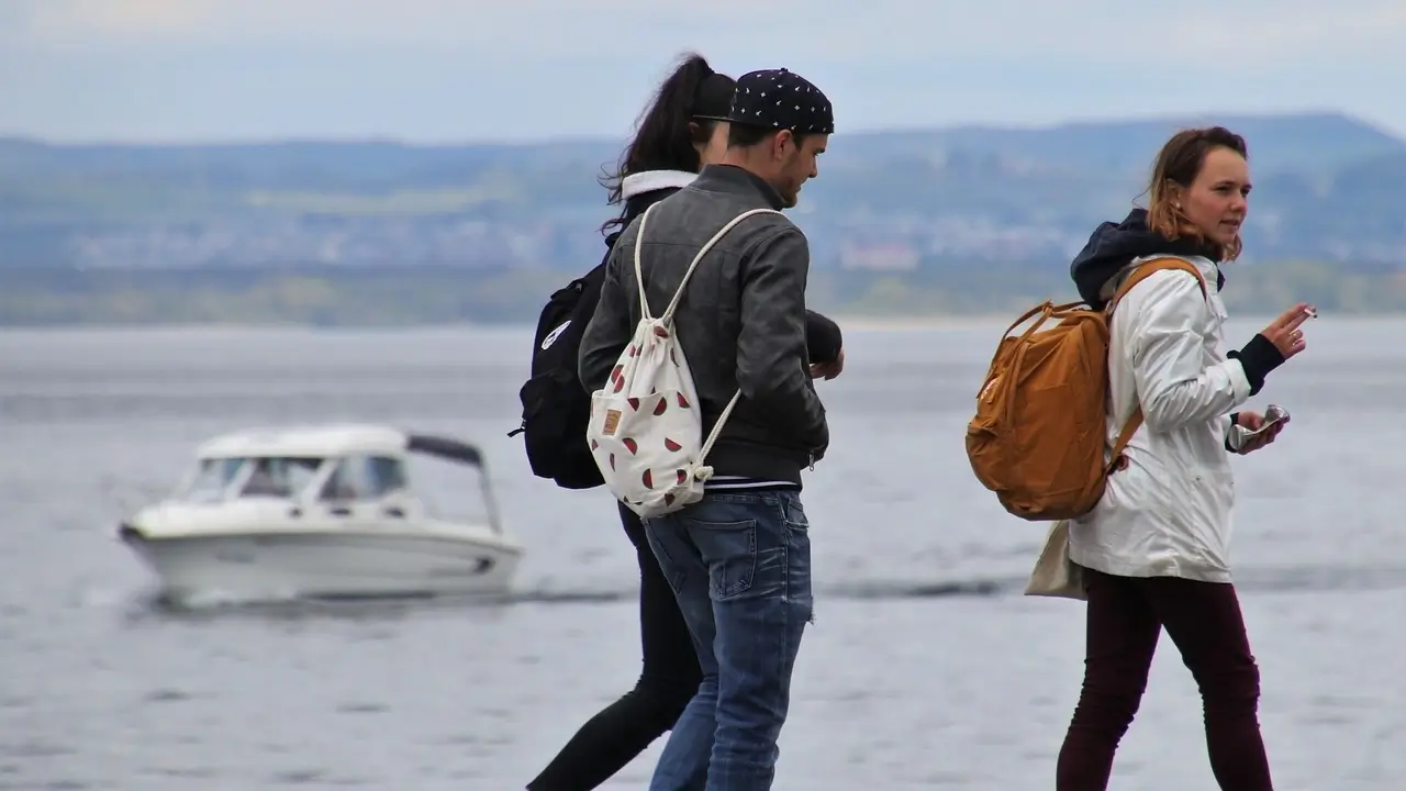 Kiedy ośrodek terapii uzależnień jest jedynym ratunkiem? 1 Obraz przedstawia trójkę osób spacerujących wzdłuż brzegu wody. Na pierwszym planie znajdują się dwie kobiety i jeden mężczyzna. Wszystkie trzy osoby są ubrane w casualowe stroje. Mężczyzna nosi czarną czapkę z daszkiem, ciemną kurtkę i dżinsy, a na plecach ma biały plecak z nadrukiem w arbuzy. Kobieta po lewej stronie, widoczna od tyłu, ma ciemne włosy związane w kucyk, nosi czarną kurtkę i ciemny plecak. Kobieta po prawej stronie nosi jasną kurtkę i brązowy plecak. W tle widać wodę, najprawdopodobniej jezioro lub morze, a także białą motorówkę na wodzie. W oddali znajdują się zielone wzgórza i niewyraźne zarysy budynków. Cała scena ma spokojny i relaksujący charakter, sugerujący wypoczynek na świeżym powietrzu.