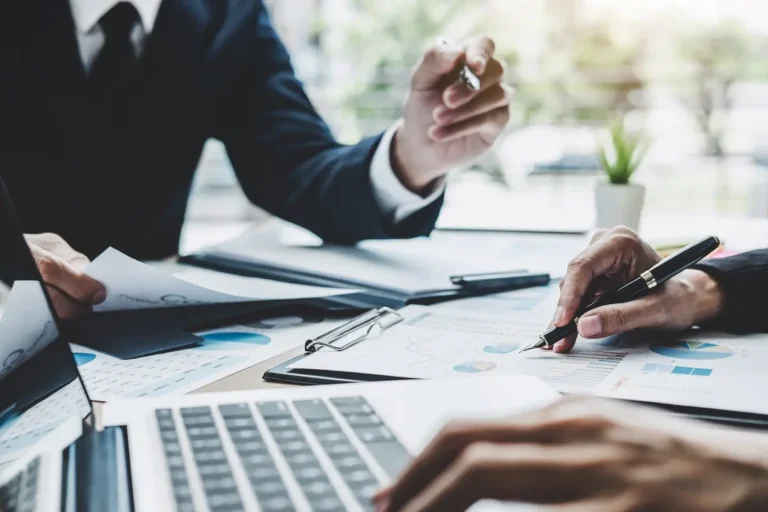 Obraz przedstawia grupę osób, prawdopodobnie w trakcie spotkania biznesowego. W centrum kadru znajdują się dwie pary rąk, z których jedna trzyma długopis i wskazuje na dokumenty leżące na stole. Dokumenty te zawierają wykresy i dane, co sugeruje, że omawiane są kwestie finansowe lub analityczne. Na stole znajduje się również otwarty laptop, spinacze do papieru oraz inne akcesoria biurowe. W tle można dostrzec rozmyte okno z widokiem na zieleń, co wskazuje na to, że spotkanie odbywa się w biurze z dostępem do światła dziennego.