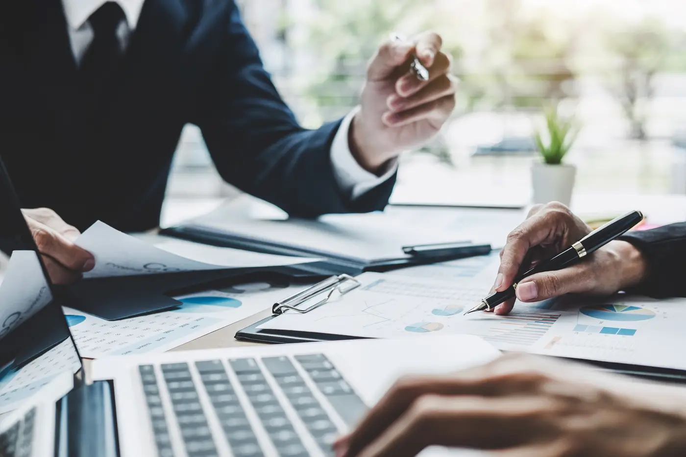 Zerkamy do statystyk, jakie są dominujące formy zatrudnienia w Polsce w 2024 roku? 1 Obraz przedstawia grupę osób, prawdopodobnie w trakcie spotkania biznesowego. W centrum kadru znajdują się dwie pary rąk, z których jedna trzyma długopis i wskazuje na dokumenty leżące na stole. Dokumenty te zawierają wykresy i dane, co sugeruje, że omawiane są kwestie finansowe lub analityczne. Na stole znajduje się również otwarty laptop, spinacze do papieru oraz inne akcesoria biurowe. W tle można dostrzec rozmyte okno z widokiem na zieleń, co wskazuje na to, że spotkanie odbywa się w biurze z dostępem do światła dziennego.