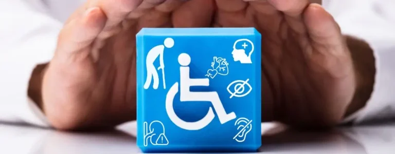 Obrazek wyróżniający do artykułu - rodzaje niepełnosprawności. Obraz przedstawia niebieski sześcian, na którym znajdują się białe ikony symbolizujące różne rodzaje niepełnosprawności. Sześcian jest otoczony dłońmi, które sprawiają wrażenie, jakby chroniły go lub otaczały opieką. Na sześcianie znajdują się następujące ikony: Ikona osoby starszej z laską, symbolizująca niepełnosprawność związana z wiekiem. Ikona wózka inwalidzkiego, symbolizująca osoby z niepełnosprawnością ruchową. Ikona osoby z problemami zdrowotnymi układu oddechowego. Ikona mózgu, symbolizująca niepełnosprawność intelektualną lub zdrowotne problemy psychiczne. Ikona oka z przekreśleniem, symbolizująca niepełnosprawność wzrokową. Ikona ucha z przekreśleniem, symbolizująca niepełnosprawność słuchową. Dłonie otaczające sześcian są rozstawione w taki sposób, jakby miały chronić sześcian, co sugeruje troskę, opiekę i wsparcie dla osób z niepełnosprawnościami. Tło obrazu jest rozmyte, co sprawia, że sześcian i dłonie są głównym punktem zainteresowania.