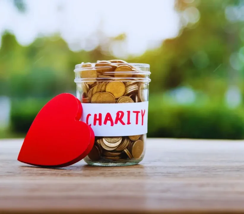 Obrazek wyróżniający do artykułu - sposoby prowadzenia zbiórek publicznych. Obraz przedstawia słoik wypełniony monetami, oznaczony napisem "CHARITY". Obok słoika znajduje się duże, czerwone, drewniane serce. Szczegóły obrazu: Słoik: Jest przezroczysty, wykonany ze szkła. Wewnątrz słoika znajdują się złote monety. Na słoiku przyklejona jest biała etykieta z ręcznie napisanym słowem "CHARITY" czerwoną farbą lub markerem. Czerwone serce: Znajduje się obok słoika, po lewej stronie. Jest duże, czerwone i wykonane z drewna lub innego twardego materiału. Tło: Tło jest rozmyte, z zielonymi i brązowymi odcieniami, co sugeruje, że zdjęcie zostało zrobione na zewnątrz, w parku lub ogrodzie. Na pierwszym planie znajduje się drewniany stół lub blat, na którym stoi słoik i leży serce. Styl: Obraz ma ciepłe, naturalne oświetlenie, co nadaje mu przyjazny i zachęcający wygląd. Kolory są żywe i kontrastowe, szczególnie czerwone serce na tle złotych monet i zielonego tła. Kontekst: Obraz jest symboliczny, nawiązujący do działalności charytatywnej i dobroczynności. Może być używany w kampaniach promujących zbiórki charytatywne, darowizny i inne formy wsparcia finansowego.