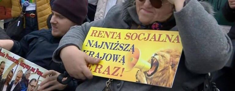 Zdjęcie do artykułu - podwyżka renty socjalnej. Na zdjęciu widzimy grupę protestujących osób, które wyrażają swoje żądania podniesienia renty socjalnej do poziomu najniższej krajowej. Opis głównego elementu zdjęcia: Osoba na pierwszym planie: Kobieta w okularach przeciwsłonecznych i szarej kurtce trzyma w rękach transparent. Transparent: Jest koloru żółtego z dużymi czerwonymi i czarnymi literami, na którym widnieje napis "RENTA SOCJALNA NAJNIŻSZĄ KRAJOWĄ TERAZ!". Obok tekstu znajduje się ilustracja ryczącego lwa z megafonem w pysku, symbolizująca siłę i determinację protestujących. Osoba w tle: W tle widoczny jest mężczyzna w ciemnej kurtce i czapce, również trzymający transparent, na którym znajdują się zdjęcia kilku osób (prawdopodobnie polityków) z napisem sugerującym ich działania lub decyzje. Protest odbywa się na zewnątrz, a uczestnicy są ubrani w ciepłe ubrania, co sugeruje chłodniejszą porę roku. Na zdjęciu widzimy grupę protestujących osób, które wyrażają swoje żądania podniesienia renty socjalnej do poziomu najniższej krajowej. Opis głównego elementu zdjęcia: Osoba na pierwszym planie: Kobieta w okularach przeciwsłonecznych i szarej kurtce trzyma w rękach transparent. Transparent: Jest koloru żółtego z dużymi czerwonymi i czarnymi literami, na którym widnieje napis "RENTA SOCJALNA NAJNIŻSZĄ KRAJOWĄ TERAZ!". Obok tekstu znajduje się ilustracja ryczącego lwa z megafonem w pysku, symbolizująca siłę i determinację protestujących. Osoba w tle: W tle widoczny jest mężczyzna w ciemnej kurtce i czapce, również trzymający transparent, na którym znajdują się zdjęcia kilku osób (prawdopodobnie polityków) z napisem sugerującym ich działania lub decyzje. Protest odbywa się na zewnątrz, a uczestnicy są ubrani w ciepłe ubrania, co sugeruje chłodniejszą porę roku.