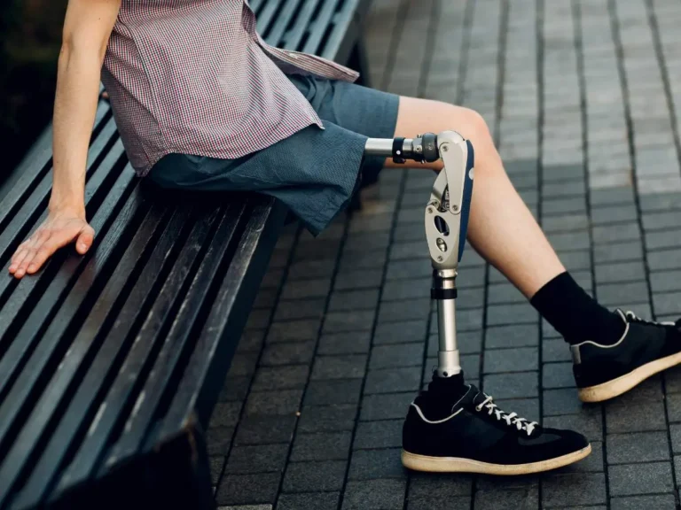 Na zdjęciu widoczna jest osoba siedząca na drewnianej ławce na zewnątrz. Osoba ta ma protezę nogi od kolana w dół. Widoczna jest lewa strona ciała osoby, która nosi szare krótkie spodenki i ciemne buty sportowe. Proteza nogi jest nowoczesna, wykonana z metalu i tworzyw sztucznych, z przegubem na wysokości kolana. Osoba opiera się jedną ręką na ławce, a nogi są rozłożone. Otoczenie sugeruje, że zdjęcie zostało zrobione na zewnątrz, być może w parku lub na deptaku, z brukowanym chodnikiem w tle.
