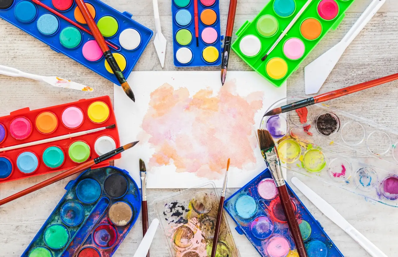 Na obrazie widzimy kolorową kompozycję zestawu farb akwarelowych, pędzli i innych narzędzi malarskich. Na środku znajduje się kartka papieru z rozpoczętym malunkiem, na którym widać pastelowe plamy w odcieniach różu, pomarańczu i żółci. Wokół tej kartki rozmieszczone są palety z farbami akwarelowymi, w różnych kolorach. Niektóre palety są czerwone, inne zielone lub niebieskie. W paletach znajdują się intensywnie kolorowe farby w różnych odcieniach – od różu, przez żółć, zieleń, po niebieski. Na paletach leżą pędzle z drewnianymi rączkami, a także plastikowe szpatułki, które są używane do mieszania farb.