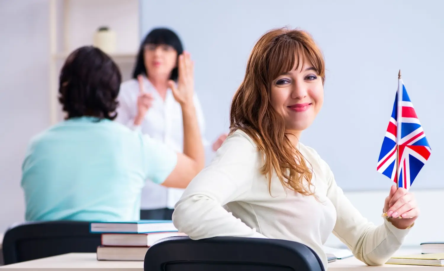 Jak umiejętność posługiwania się angielskim wpływa na ścieżkę zawodową? 1 Na zdjęciu znajduje się młoda kobieta, która siedzi przy biurku i trzyma w dłoni małą flagę Wielkiej Brytanii. Kobieta jest uśmiechnięta i patrzy prosto w obiektyw aparatu. W tle widać dwie inne osoby — jedna z nich siedzi przy stole, a druga stoi i gestykuluje, prawdopodobnie coś wyjaśniając. Zdjęcie może sugerować kontekst związany z nauką języka angielskiego lub pracą w środowisku międzynarodowym.