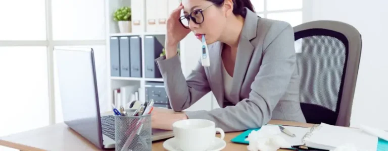 Obrazek do artykułu - czy można zwolnić pracownika na L4. Obraz przedstawia kobietę siedzącą przy biurku w biurze, która wygląda na chorą. Ubrana jest w szary elegancki garnitur, nosi okulary i wydaje się zmęczona. Kobieta ma w ustach termometr, co sugeruje, że może mieć gorączkę. Opiera głowę na dłoni, co podkreśla jej wyczerpanie. Na biurku przed nią znajduje się otwarty laptop, kubek z kawą, koszyk z długopisami i inne biurowe przedmioty, takie jak notes i kilka zgniecionych chusteczek, co dodatkowo podkreśla stan jej zdrowia. Wygląda na to, że mimo złego samopoczucia, kobieta pracuje.