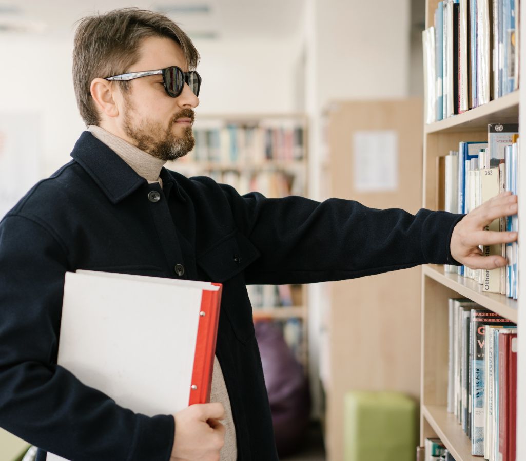 Praktyczny przewodnik dla pracodawcy o tym, jaką pracę może wykonywać osoba niewidoma 1 Obrazek do artykułu - jaką pracę mogą wykonywać osoby niewidome. Obraz przedstawia niewidomego mężczyznę w okularach przeglądającego książki na półce. Mężczyzna ma ciemne włosy i brodę, ubrany jest w ciemny płaszcz i trzyma pod pachą duży segregator w kolorze białym z czerwonym grzbietem. Jego prawa ręka jest wyciągnięta w stronę półki z książkami, a palce dotykają jednej z książek, jakby szukał czegoś konkretnego. W tle widać rozmyte regały z książkami, co wskazuje, że scena rozgrywa się w biurze, bibliotece lub czytelni. Atmosfera na obrazie jest spokojna, sugerująca koncentrację na wykonywanej czynności.