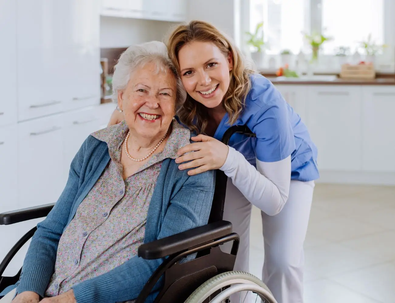 Rozwiewamy wątpliwości, komu nie przysługuje świadczenie wspierające 1 Na obrazie widzimy starszą kobietę siedzącą na wózku inwalidzkim, uśmiechniętą i pogodną. Jest ubrana w jasną bluzkę z delikatnym wzorem kwiatowym oraz niebieski sweter. Na jej szyi widać perłowy naszyjnik. Za nią stoi młoda kobieta w niebieskim uniformie medycznym, również uśmiechnięta, obejmująca ramiona starszej pani w przyjaznym, troskliwym geście. Kobieta wygląda na opiekunkę lub pielęgniarkę. Scena jest jasna, z przytulnym, domowym wnętrzem w tle, co nadaje obrazowi pozytywny i ciepły wydźwięk.