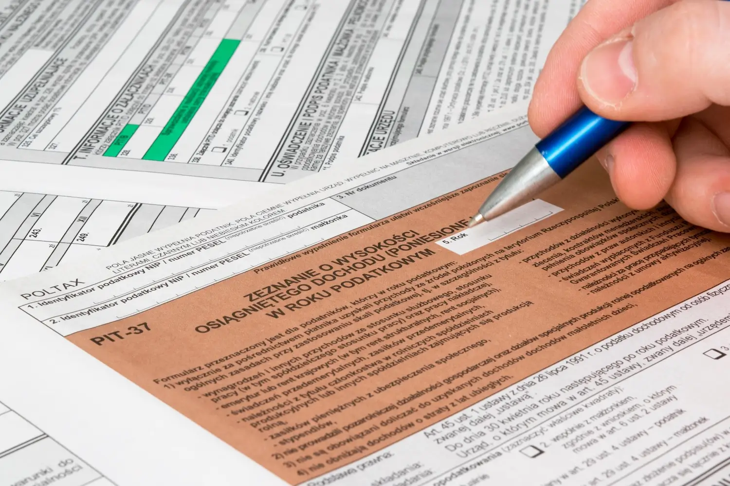 Obraz przedstawia zbliżenie na rękę osoby, która wypełnia formularz PIT-37, polski dokument do rozliczenia podatkowego. Na pierwszym planie widoczny jest fragment wypełnianego formularza z napisem „Zeznanie o wysokości osiągniętego dochodu (poniesionej straty) w roku podatkowym”. Osoba trzyma długopis i wypełnia odpowiednie pola. W tle znajdują się inne formularze podatkowe, co sugeruje, że to scena związana z rozliczaniem podatków. Kolorystyka formularzy to odcienie brązu, zieleni i bieli.