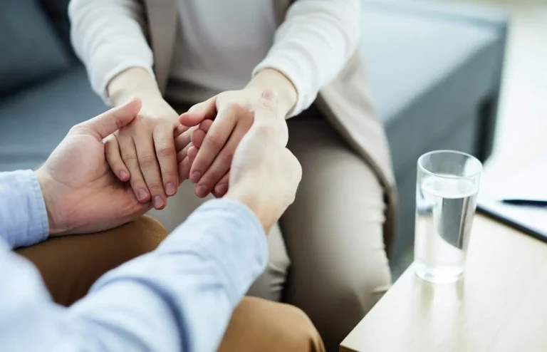 Obraz przedstawia zbliżenie na dwie osoby siedzące naprzeciw siebie, trzymające się za ręce. Gest ten sugeruje wsparcie emocjonalne lub próbę pocieszenia. Osoba po lewej stronie, w jasnoniebieskiej koszuli, trzyma dłoń drugiej osoby, ubranej w beżowe ubrania. W prawym dolnym rogu, na stole, stoi szklanka wody. Tło jest rozmyte, z widoczną częścią szarej sofy, co tworzy spokojną, intymną atmosferę. Obraz emanuje poczuciem bliskości, zrozumienia i wsparcia, co sugeruje rozmowę o trudnych emocjach lub moment wsparcia psychologicznego.