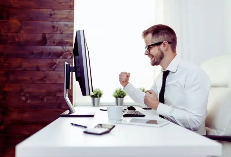 Obrazek do artykułu - dlaczego warto zatrudniać niewidomych do tworzenia treści. Na zdjęciu widzimy mężczyznę w grubych okularach w średnim wieku, siedzącego przy biurku przed komputerem. Mężczyzna jest ubrany w białą koszulę i krawat, co wskazuje na formalny ubiór biurowy. Wygląda na bardzo zadowolonego lub podekscytowanego – na jego twarzy widoczny jest szeroki uśmiech, a dłonie są zaciśnięte w geście zwycięstwa, jakby właśnie osiągnął coś ważnego. Na biurku przed nim znajdują się różne przedmioty, takie jak filiżanka, kalkulator, długopis oraz notatnik, co sugeruje, że mężczyzna wykonuje pracę biurową. Tło zdjęcia jest dość minimalistyczne – z prawej strony widać drewnianą ścianę, co dodaje wnętrzu ciepłego i przyjemnego charakteru. Cała scena oddaje atmosferę sukcesu i zadowolenia z osiągnięcia celów w pracy.