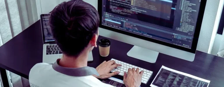 Obrazek do artykułu - dlaczego warto zatrudniać osoby niewidome w branży IT? Na zdjęciu widzimy osobę siedzącą przy biurku i pracującą na komputerze. Osoba jest widoczna od tyłu, co pozwala nam dostrzec jej skupienie na ekranie monitora. Przed monitorem znajduje się duży komputer stacjonarny, na ekranie którego widoczny jest otwarty program z kodem, sugerujący, że osoba ta zajmuje się programowaniem lub pracą związaną z IT. Osoba ma krótkie, ciemne włosy, a ubrana jest w jasną koszulę. Na biurku obok komputera znajduje się laptop, kubek na kawę oraz dokumenty z wydrukowanym tekstem, który również wygląda jak kod lub techniczne informacje. Pomieszczenie jest dobrze oświetlone, a zasłony na oknie po lewej stronie dodają lekkości całemu wnętrzu. Atmosfera zdjęcia sugeruje intensywną pracę i skupienie, typowe dla osoby zajmującej się programowaniem lub innymi zadaniami wymagającymi dużej uwagi do szczegółów.