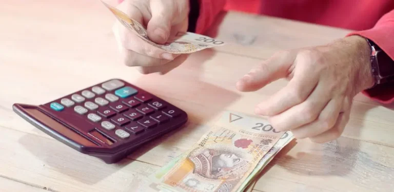 Obrazek do artykułu - ile pieniędzy należy się osobie z grupą inwalidzką. Na obrazie widać osobę przeliczającą polskie banknoty o nominale 200 złotych. Osoba trzyma banknoty w obu dłoniach. Na stole, po lewej stronie, znajduje się ciemnoczerwona kalkulator z dużymi, widocznymi przyciskami, a obok niego kilka banknotów. Osoba nosi czerwony strój, co stanowi kontrast z jaśniejszym drewnianym tłem stołu. Scena sugeruje czynność przeliczania lub zarządzania pieniędzmi.