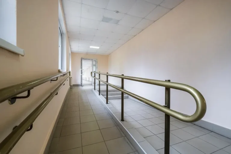 Na zdjęciu znajduje się korytarz wewnętrzny z zamontowanymi poręczami po obu stronach, które są przeznaczone do ułatwienia poruszania się osobom niepełnosprawnym. Podłoga jest wyłożona jasnoszarymi płytkami, a ściany są pomalowane na pastelowy odcień beżu. Poręcze mają metaliczny, złoty kolor i biegną równolegle wzdłuż rampy prowadzącej do drzwi na końcu korytarza. Całe pomieszczenie jest jasno oświetlone przez lampy sufitowe, a po lewej stronie widać duże okno, które wpuszcza naturalne światło.